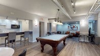 Located in Dayton, Ohio, this single-floor entertainment space is thoughtfully designed to accommodate socializing, gaming, and relaxed gatherings. The open layout flows seamlessly between distinct functional zones, all unified by durable wood-look flooring and a neutral gray-and-white color palette.

The centerpiece of the space is a full-size, ornately carved wooden pool table positioned beneath a decorative Tiffany-style pendant light. Exposed ductwork and industrial-style track lighting run overhead, lending the area an urban, loft-inspired character. A wall-mounted cue rack nearby keeps the gaming area organized and functional.

Adjacent to the billiards zone, a comfortable lounge area features a cognac leather sofa, leather accent chairs, and a glass-top foosball table, creating an inviting spot for casual conversation or game play. Colorful abstract artwork adorns the walls throughout, adding visual energy to the space.

A dedicated seating alcove with a cinema-themed décor and floor lamp provides a more intimate retreat, while high-top tables and bar stools offer additional gathering spots. The compact kitchenette is well-appointed with white shaker cabinetry, a quartz countertop, subway tile backsplash, stainless steel sink, and a black refrigerator, making refreshments readily accessible for guests.