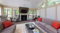 Located at 3151 Exacta Dr in Raleigh, NC, this single-floor space is designed to impress with its open, airy layout and thoughtful architectural details. The main living area showcases dramatic vaulted ceilings that rise to striking heights, lined with a wall of large-pane windows that flood the interior with natural light and frame lush, wooded outdoor views. A classic fireplace with a white mantel and dark tile surround anchors one end of the room, complemented by a bold accent wall that adds a contemporary touch. The generous layout accommodates both a comfortable lounge seating area and a casual dining space, making it highly versatile for gatherings and everyday use. Plush carpeting in a warm neutral tone runs throughout, adding comfort underfoot, while a patterned area rug defines the central seating zone. An adjacent area features soaring double-height ceilings supported by elegant columns, further emphasizing the grandeur of the space. A built-in kitchenette or serving area with white cabinetry is visible nearby, enhancing the functionality of the space. Throughout, recessed lighting and an abundance of natural light create a warm, welcoming ambiance suited for community or social use.