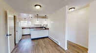 Located at 17840 32nd Avenue South in SeaTac, Washington, Unit V63 is a single-floor residence thoughtfully laid out for comfortable everyday living. The open living area features wood-style luxury vinyl flooring, a textured ceiling, and a striking white-painted brick fireplace that serves as a natural focal point. Baseboard heating runs throughout, providing efficient warmth across the space. The kitchen is well-appointed with white cabinetry, dark countertops, a peninsula with pendant lighting overhead, a full suite of white appliances including a refrigerator, electric range, and dishwasher, a window above the sink for natural light, and an adjacent open pantry for additional storage. The bedroom is carpeted for comfort and includes a reach-in closet with built-in rod and shelving. The bathroom features wood-style flooring, a vanity with a dark countertop, and a full tub-shower combination with white tile surround. Throughout the unit, clean white walls and consistent finishes create a bright, cohesive aesthetic. The layout flows naturally from the entryway through the living and dining areas into the kitchen, making the space feel open and functional.