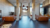 Located at 3000 Fast Ln in Fort Mill, SC, this contemporary lobby space makes an immediate impression with its dramatic double-height ceilings and expansive floor-to-ceiling windows that flood the interior with natural light. The entrance is framed by substantial concrete columns and a bold glass curtain wall facade, lending an industrial-modern aesthetic to the overall design.

Dark slate tile flooring extends throughout the main corridor and lounge areas, providing a sleek, cohesive foundation. The space is organized into distinct seating zones, each thoughtfully curated with a mix of cognac leather sectional sofas, white upholstered chaises, and accent chairs arranged atop layered area rugs. Sculptural pendant lighting in varied shapes hangs from the soaring ceiling, complementing warm wood slat ceiling panels in select areas.

A standout feature is a large decorative lattice-style wall partition in natural wood tones, adding architectural texture and visual depth. Bold abstract artwork displayed against a deep charcoal accent wall creates a gallery-like atmosphere in one of the lounge alcoves, accompanied by a large sculptural birch branch installation in a planter. A dining area with a dark table and mixed seating offers a functional gathering space. The overall ambiance is sophisticated, warm, and welcoming.