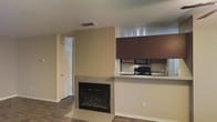 This single-floor residence offers an open, functional layout that seamlessly connects the living and kitchen areas. The living space is anchored by a fireplace framed in neutral tile, adding a focal point to the room while complementing the warm-toned, dark hardwood flooring that extends throughout. The walls are finished in a soft beige tone, contributing to a clean and understated aesthetic.

The kitchen is positioned adjacent to the living area, separated by a raised countertop that doubles as a breakfast bar or pass-through surface. Dark reddish-brown cabinetry overhead provides ample storage, and the space is equipped with black appliances including a gas range and built-in microwave. A single-basin sink sits along the counter near the pass-through, making the kitchen efficient and accessible.

A hallway visible from the main living area leads to additional rooms, suggesting a practical and well-organized floor plan. A standard door opens to what appears to be a closet or additional room, adding to the overall functionality of the space. The layout is straightforward, with recessed ceiling vents ensuring consistent climate control throughout.