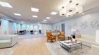 Located at 2012 N Orange Ave in Sarasota, Florida, this thoughtfully designed single-floor community space offers a generous, open layout across a well-appointed footprint. The interior presents a clean, contemporary aesthetic defined by light-toned flooring, soft neutral walls, and an abundance of natural light streaming through large windows dressed with white blinds and sheer curtains.

The space flows seamlessly from a welcoming lounge area near the entrance — furnished with plush white sofas, warm cognac leather armchairs, and a glass coffee table atop an area rug — into a central reception zone featuring a sleek white desk with modern blue task chairs and curated wall art. Recessed skylights and panel lighting illuminate the open corridor, which connects the various functional areas.

A dedicated dining area accommodates multiple tables and chairs beneath additional skylight panels, with expansive windows offering views of lush outdoor greenery — creating a bright, airy atmosphere ideal for gatherings. Adjacent to the dining space is a fully equipped kitchen featuring a large island with bar seating, pendant lighting, a stainless steel refrigerator, and a ventilated range — all finished with a textured stone backsplash. A built-in salon station with a styling chair, vanity mirror, and product display rounds out this versatile, amenity-rich space.