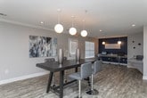 Located at 150 Orchard Brook Dr in Gainesville, Georgia, this single-floor communal space offers a thoughtfully designed, open-concept layout ideal for gatherings and everyday resident use. The interior is finished with warm wood-look luxury vinyl plank flooring throughout, crisp white walls with neutral gray accents, and crown molding that lends a polished, cohesive aesthetic.

The main lounge area features a ceiling fan, recessed lighting, and a wall-mounted flat-screen TV, creating a comfortable setting for relaxation. Adjacent to the lounge is a fully equipped kitchenette outfitted with gray cabinetry, stone tile backsplash, stainless steel appliances including a full-size refrigerator and microwave, and a dedicated counter workspace. A high-top bar table with bar stools positioned near a large window completes the casual dining setup, allowing for natural light to fill the space.

A defined dining area hosts a bold, dark high-top table with sleek chrome-accented seating, illuminated by a trio of globe pendant lights. Beyond, a dedicated office or business center area offers workstations framed by additional natural light from large windows. Throughout the space, subtle tray ceiling details add architectural dimension, while recessed lighting ensures bright, even illumination. Multiple access points and clearly marked exits make the space highly functional and accommodating.