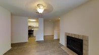 This single-story space offers an open-concept layout that seamlessly connects the living area to the kitchen. The living room is anchored by a tiled fireplace with a sleek black surround, providing a focal point that adds warmth and character to the space. Hardwood-style flooring runs throughout, lending a clean, cohesive aesthetic across the main areas. A ceiling fan with a light fixture is centrally positioned, offering both ventilation and ambient lighting.

The kitchen is visible from the living area, separated by a partial wall that maintains an open feel while defining each zone. It features white cabinetry, light countertops, and stainless steel appliances, including a gas range and overhead ventilation hood. The kitchen benefits from its own dedicated lighting, enhancing functionality. Neutral white walls throughout the space create a bright, versatile backdrop. A partially visible adjoining area, framed by columns and a half-wall with decorative molding, suggests additional functional space, potentially a dining area. The overall ambiance is clean, functional, and move-in ready.