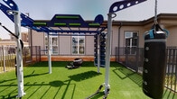 Located at Bellaire Boulevard in Richmond, Texas, this single-story amenity clubhouse is designed to offer residents a well-rounded recreational experience. The interior centers around a vibrant game room, where a full-sized pool table commands attention beneath a cascading pendant light fixture. The room is defined by a bold navy accent wall contrasted with a striking blue and gold geometric patterned wallcovering, creating a lively yet sophisticated atmosphere. Arcade machines and a wall-mounted flat-screen television round out the entertainment options, while large multi-pane windows flood the space with natural light and provide views of the surrounding community. Comfortable built-in lounge seating with upholstered cushions and curated wall art offer relaxed gathering spots, complemented by warm hardwood-style flooring throughout. The outdoor area extends the amenity experience with a dedicated fitness zone featuring synthetic turf, a multi-function overhead rig with monkey bars and climbing attachments, battle ropes, and a heavy tire for functional training. The building's exterior reflects a classic architectural style with neutral siding and white-trimmed windows, blending seamlessly into the neighborhood setting. Together, the indoor and outdoor spaces create a cohesive, activity-focused environment suited for both social and fitness-oriented use.