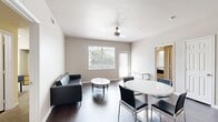 This well-designed 5-bedroom, 4-bathroom unit offers a functional and comfortable layout across a single floor. The open living and dining area serves as the heart of the home, featuring dark hardwood flooring, neutral walls, and ample natural light from large windows. A ceiling fan adds comfort to the shared space, which comfortably accommodates both a seating area and a dining table. The galley-style kitchen is efficiently organized with light wood cabinetry, dark granite-style countertops, a double sink, and white appliances including a refrigerator, stove, and built-in microwave. Each bedroom is carpeted and includes a ceiling fan, a window with blinds for natural light and privacy, a built-in desk, and a mirrored sliding-door closet — providing each resident with a private, functional retreat. The four bathrooms are consistently finished with wood-look flooring, light wood vanity cabinets, dark countertops, and tiled shower-tub combinations featuring a classic black accent tile border. A central hallway connects all rooms, contributing to an efficient and well-organized flow throughout the unit. This space is well-suited for multi-resident living, combining shared common areas with private individual accommodations.