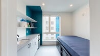 This single-floor unit is thoughtfully designed to maximize functionality within a compact footprint. The main living area combines sleeping and kitchen spaces seamlessly, featuring a platform bed with built-in drawer storage beneath, and a wall-mounted mini-split air conditioning unit for individual climate control. Recessed ceiling lighting keeps the space feeling bright and open.

The kitchenette is positioned along one wall, offering a granite-speckled countertop, white cabinetry with black hardware, a compact refrigerator, and open shelving above that houses a microwave. A large window adjacent to the kitchenette invites generous natural light and provides views of the surrounding urban building exteriors. A bold teal accent wall adds a striking pop of color, contrasting cleanly against the otherwise crisp white walls and light flooring.

The entryway features a smart panel control system, a full-length mirror, and access to an open closet with built-in shelving. The private bathroom is clean and minimal, finished in white with black matte grab bars and fixtures, offering both practicality and a modern aesthetic. The overall design is streamlined and contemporary, making efficient use of every corner while maintaining a fresh, uncluttered atmosphere throughout.