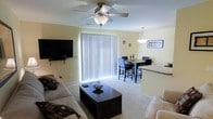 This single-floor space features an open-concept design that seamlessly connects the living and dining areas, creating a cohesive and functional layout. The living area is anchored by a plush sofa set arranged around a dark wood coffee table, with a wall-mounted flat-screen television serving as a focal point. A ceiling fan with an integrated light fixture provides both ventilation and ambient lighting overhead.

Large vertical blinds covering a full-length sliding glass door or oversized window allow for adjustable natural light while offering privacy. The neutral, light yellow walls and wall-to-wall carpeting contribute to a warm and inviting atmosphere throughout the space.

Adjacent to the living area, the dining space accommodates a dark wood table with matching chairs, illuminated by a pendant light fixture that adds a refined touch. A partial view of what appears to be a kitchen counter with a light-colored surface is visible nearby, suggesting a convenient eat-in or bar-style transition between the kitchen and dining areas. Decorative wall art accents the dining space, adding character to the overall interior.