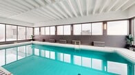 Located at 1975 Boulevard de Maisonneuve Ouest in Montréal, this indoor pool facility offers a well-appointed aquatic space designed for both leisure and lap swimming. The centerpiece is a generously sized rectangular pool with vibrant turquoise water, framed by clean white mosaic tile surrounds that extend along the pool deck.

The space is defined by a striking structural ceiling featuring a grid of exposed white ribs, adding an architectural rhythm overhead while contributing to the room's bright, airy character. Walls are finished in a combination of crisp white and warm taupe tones, creating a calm and modern atmosphere.

Natural light pours in through large floor-to-ceiling windows and glass sliding doors that line one side of the pool, offering views of an adjacent outdoor rooftop terrace equipped with lounge chairs and a picnic table — accessible directly from the pool area. This seamless indoor-outdoor connection enhances the sense of openness and elevates the overall experience of the space.

Along the poolside, wall-mounted sconce lighting and a series of modern seating chairs provide comfortable poolside areas for relaxation. The facility's elevated position within the building affords views of the surrounding Montréal cityscape through the windows, adding an urban backdrop to the serene aquatic environment.