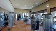 This fitness center is a thoughtfully designed, single-floor facility built to support a comprehensive workout experience. The space is generously proportioned, with elevated ceilings that create an open, airy atmosphere and prevent any sense of overcrowding even when fully equipped with machines. Recessed lighting throughout the ceiling ensures bright, even illumination across the entire floor, complemented by ample natural light streaming in through multiple large, dark-framed windows lining the walls. The flooring features a warm hardwood-style surface that adds a polished, welcoming aesthetic while providing a durable workout surface. The equipment lineup includes an impressive array of Precor-brand strength training machines and cardiovascular equipment, such as ellipticals and cable machines, catering to a full range of fitness goals. A flat-screen TV is mounted near the ceiling for entertainment during workouts, and a vending machine is conveniently positioned near the entrance. Full-length mirrors are integrated along one wall, enhancing the sense of space and allowing users to monitor their form. The overall ambiance is clean, modern, and professional, making this fitness center a functional and inviting space for all levels of fitness enthusiasts.