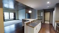 This single-floor space showcases a thoughtfully designed open-concept layout that seamlessly connects the kitchen and living areas. The kitchen serves as the focal point, anchored by a substantial island featuring dual sinks, white cabinetry with gold-toned trim detail, and a mosaic tile accent strip. A modern pendant light hangs above, providing task lighting and a contemporary design element. The countertops offer generous workspace, complemented by additional cabinetry and open shelving along the perimeter wall, providing ample storage solutions.

The flooring transitions from rich, dark hardwood in the kitchen and adjacent areas to carpeting in the living space, creating a natural visual distinction between zones while maintaining a cohesive aesthetic. The ceiling design is particularly notable, featuring a dramatic exposed concrete panel that adds an industrial character to one section, contrasting with the standard white ceiling in adjacent areas. Neutral gray walls throughout provide a versatile backdrop. The living area benefits from natural light through a window fitted with dark curtains, while doorways visible in the background suggest additional rooms beyond the main open space.