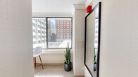 This thoughtfully designed studio suite offers an efficient yet stylish urban living experience across a single floor. The open layout maximizes every inch of space, seamlessly integrating the sleeping, living, and kitchen areas into a cohesive, functional environment.

The sleeping area serves as the centerpiece of the suite, anchored by a striking curved canopy bed frame that adds a sculptural, modern aesthetic. Large windows wrap around the space, flooding the room with abundant natural light and framing impressive panoramic views of the surrounding cityscape, including neighboring mid-rise buildings and urban rooftops.

A dedicated living area features a central column with a wall-mounted television, alongside a teal accent chair and a bistro-style high table with seating — ideal for relaxing or working. The neutral carpeted flooring and cream-toned walls create a calm, contemporary backdrop throughout.

The kitchenette is well-equipped with dark espresso cabinetry, open glass-front upper shelving, a stainless steel refrigerator, microwave, cooktop, and a sink — providing all the essentials for convenient in-suite dining. A compact entry corridor with a large mirror and additional storage rounds out the layout. Lush indoor plants are thoughtfully placed throughout, adding a refreshing natural element to the urban setting.