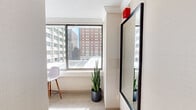 This thoughtfully designed studio suite offers an efficient yet stylish urban living experience across a single floor. The open layout maximizes every inch of space, seamlessly integrating the sleeping, living, and kitchen areas into a cohesive, functional environment.

The sleeping area serves as the centerpiece of the suite, anchored by a striking curved canopy bed frame that adds a sculptural, modern aesthetic. Large windows wrap around the space, flooding the room with abundant natural light and framing impressive panoramic views of the surrounding cityscape, including neighboring mid-rise buildings and urban rooftops.

A dedicated living area features a central column with a wall-mounted television, alongside a teal accent chair and a bistro-style high table with seating — ideal for relaxing or working. The neutral carpeted flooring and cream-toned walls create a calm, contemporary backdrop throughout.

The kitchenette is well-equipped with dark espresso cabinetry, open glass-front upper shelving, a stainless steel refrigerator, microwave, cooktop, and a sink — providing all the essentials for convenient in-suite dining. A compact entry corridor with a large mirror and additional storage rounds out the layout. Lush indoor plants are thoughtfully placed throughout, adding a refreshing natural element to the urban setting.