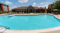 This single-story community facility is defined by its expansive outdoor swimming pool, which serves as the centerpiece of the property. The pool is generously sized with clean, clear blue water and a classic rectangular shape with curved corners, surrounded by a wide concrete pool deck. Several lounge chairs are arranged along one side, providing ample space for relaxation and sunbathing under open skies.

The building itself features a traditional architectural style with sage green horizontal siding, a steeply pitched dark brown shingled roof, and a prominent central entrance framed by large multi-pane windows that allow abundant natural light into the interior. Decorative circular detailing accents the roofline above the entrance, adding a refined touch to the overall design. Potted plants and greenery line the front of the building, softening the transition between the structure and the pool deck.

An adjacent brick structure is partially visible to the right, suggesting additional facilities on the property. The overall ambiance is bright, open, and resort-like, with mature trees visible in the background contributing to a landscaped, welcoming environment.