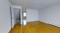 This well-maintained space spans three floors and presents a clean, straightforward design with a welcoming ambiance. The interior is characterized by polished hardwood flooring in a warm honey tone that runs throughout, complementing the crisp white walls and ceilings that give each room a bright, airy feel. Natural light filters in through windows, enhancing the open and uncluttered atmosphere. The layout allows for a smooth flow between areas, with clear sightlines from room to room. A doorway leads into an adjacent space that benefits from additional natural light through a front-facing window, suggesting a versatile entry or transitional area. The clean architectural lines and neutral palette provide an excellent blank canvas, making the space highly adaptable for a variety of uses or personal styles. Baseboard heating elements are present, ensuring year-round comfort. The multiple floors offer distinct zones for different functions, adding depth and versatility to the overall layout. The overall condition of the space reflects careful upkeep, with well-finished surfaces and a tidy, move-in ready presentation throughout.