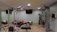 This single-level fitness space is thoughtfully designed to accommodate a variety of exercise routines. The room features light sage-green walls that create a calm, focused atmosphere, complemented by wood-look flooring that adds warmth to the functional environment. Recessed ceiling lights provide consistent, even illumination throughout the space.

The room is equipped with a multi-function cable machine ideal for strength training, along with a dedicated weight bench for targeted exercises. A medicine ball rack along one wall organizes an assortment of weighted balls for functional fitness training, while several exercise balls of varying sizes add versatility for core and balance workouts. Mirrored wall panels enhance the sense of space and allow users to monitor their form during exercise.

A wall-mounted flat-screen television is centrally positioned, offering entertainment or instructional content during workouts. A ventilation grille ensures proper airflow, maintaining comfort during intense training sessions. Overall, the space is efficiently laid out to maximize usability while maintaining an organized and clean aesthetic suited for serious fitness activity.