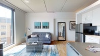 This single-floor space offers a clean, modern aesthetic defined by crisp white walls, warm hardwood flooring, and a thoughtfully designed open layout. The living area serves as the focal point, featuring a contemporary grey tufted sofa paired with metallic accent pillows and a geometric mirrored coffee table. A vibrant blue and gold abstract area rug anchors the seating arrangement, adding a pop of color that complements the two teal and gold canvas artworks displayed on the wall. A sleek arc floor lamp provides additional ambient lighting alongside the bright, evenly lit white ceiling, which features subtle panel detailing. A large full-length mirror with a bold black frame enhances the sense of depth and space. An open doorway leads to a secondary room, visible in the reflection as a kitchen or utility area with stainless steel appliances. To the right, a stainless steel refrigerator and white cabinetry hint at an adjoining kitchen space. The overall ambiance is contemporary and inviting, with a neutral base palette accented by bold décor choices that give the space a polished, well-curated feel.