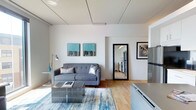 This single-floor space offers a clean, modern aesthetic defined by crisp white walls, warm hardwood flooring, and a thoughtfully designed open layout. The living area serves as the focal point, featuring a contemporary grey tufted sofa paired with metallic accent pillows and a geometric mirrored coffee table. A vibrant blue and gold abstract area rug anchors the seating arrangement, adding a pop of color that complements the two teal and gold canvas artworks displayed on the wall. A sleek arc floor lamp provides additional ambient lighting alongside the bright, evenly lit white ceiling, which features subtle panel detailing. A large full-length mirror with a bold black frame enhances the sense of depth and space. An open doorway leads to a secondary room, visible in the reflection as a kitchen or utility area with stainless steel appliances. To the right, a stainless steel refrigerator and white cabinetry hint at an adjoining kitchen space. The overall ambiance is contemporary and inviting, with a neutral base palette accented by bold décor choices that give the space a polished, well-curated feel.