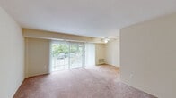 This single-floor residence offers a well-organized layout designed for comfortable everyday living. The open living area is a standout feature, highlighted by large sliding glass doors dressed with vertical blinds that frame views of lush greenery and invite abundant natural light. A ceiling fan overhead adds both comfort and function to the space, which is finished with warm, neutral-toned carpeting throughout. The layout also includes a secondary carpeted room with its own sliding glass door, providing additional flexibility as a bedroom or den.

The kitchen is efficiently designed with light wood cabinetry extending to the ceiling for generous storage, a tile-pattern floor, and white appliances including a gas range, dishwasher, and refrigerator. A peninsula counter connects the kitchen to the adjacent living space, offering a convenient pass-through and casual dining or prep area.

The bathroom is clean and functional, featuring white tile surround, a tub-shower combination, a mirrored medicine cabinet, a wood-trimmed vanity with under-sink storage, and a lighted fixture above. Storage is further addressed by a spacious walk-in closet with built-in shelving and hanging rods. Overall, the space presents a bright, neutral palette throughout.