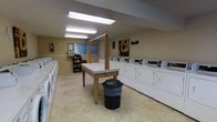 This shared laundry facility is a spacious, single-floor room designed to accommodate high-volume laundry needs for a communal setting. The room is lined on both sides with rows of white top-load washers and dryers, providing ample capacity for multiple users at once. Bright fluorescent ceiling lights illuminate the space evenly, ensuring a well-lit and functional environment throughout the day.

At the center of the room stands a large, sturdy folding table with a wooden base and a light-colored laminate surface, offering generous space for sorting and folding laundry. A vertical hanging bar is mounted above the table, providing a convenient spot for hanging garments.

Along one wall, a small bookshelf stocked with books and a chair creates a modest reading nook, allowing users to relax while waiting for their laundry. A wall clock is mounted nearby for easy time-keeping. Decorative artwork adds a touch of warmth to the otherwise utilitarian space.

The room features light yellow-painted walls and easy-to-clean vinyl flooring, reflecting a practical yet welcoming aesthetic. A window with vertical blinds allows natural light to filter in, complementing the overhead lighting. A waste bin is conveniently placed near the folding table. Overall, this laundry room is thoughtfully arranged to be both efficient and comfortable for everyday use.