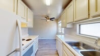 This single-story unit in Concord, California offers a straightforward, functional layout with distinct living, kitchen, and bathroom areas. The galley-style kitchen is outfitted with cream-toned cabinetry providing ample upper and lower storage, white laminate countertops, a double stainless steel sink, a coil-top electric range, and a dishwasher. A ceiling fan with integrated lighting anchors the kitchen ceiling, while a window allows natural light to filter in, contributing to a bright, airy feel. Light-toned wood-style flooring runs throughout the kitchen, adding a clean, cohesive look.

The living area features soft, neutral-toned carpeting and benefits from a large sliding glass door that opens to a private balcony, offering views of mature trees and the surrounding outdoor setting. An additional carpeted room with a ceiling fan and window provides a comfortable, well-lit space suitable as a bedroom. Another carpeted bedroom receives ample natural light through a sizable window with views of greenery outside.

The bathroom includes a white vanity with under-sink cabinetry, a broad mirror, and a well-lit vanity light bar above. Wood-style flooring continues into the bathroom, maintaining a consistent aesthetic throughout the unit. Overall, the space presents a clean, practical environment suited for everyday living.