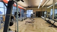 This fitness center is designed to support a comprehensive workout experience within a single open-plan floor. The space is furnished with a varied selection of exercise equipment, including elliptical trainers, treadmills, weight machines, and free weight stations with an adjustable bench, catering to both cardio and strength training needs. Large floor-to-ceiling windows line one wall, framing outdoor views and allowing abundant natural light to fill the room, creating an open and energizing atmosphere. The ceiling rises generously overhead and is fitted with ceiling fans that aid in air circulation during workouts. Overhead tube lighting supplements natural light, ensuring the space remains well-lit throughout the day. The flooring consists of light wood-toned panels that contribute a clean, modern aesthetic. An accent wall painted in a bold orange-red adds a dynamic visual contrast to the otherwise neutral color palette of beige and tan walls. A large wall-mounted mirror on one side of the room enhances the sense of space and allows users to monitor their form. A dark wood door provides access, and a small chalkboard near the entrance offers a functional touch for posting workout information or announcements.