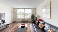 This single-floor residence offers a thoughtfully arranged layout that balances comfort and functionality across several well-defined living areas. The living room serves as the heart of the home, featuring warm hardwood-style flooring, a textured ceiling, and generous windows that frame treetop views and fill the space with natural light. A leather sofa anchored by a patterned area rug and framed wall art lend the room a relaxed, curated character. The dining area connects seamlessly to the kitchen through a pass-through opening, creating an open and communicative flow between spaces. The kitchen is efficiently designed with gray cabinetry, white countertops, tile backsplash, and a full suite of white appliances including a refrigerator, dishwasher, and electric range. The bedroom is a calm retreat, carpeted for comfort, and equipped with a large built-in closet offering ample storage. A broad window brings in natural light and exterior views. Dark wood furniture, including a dresser with mirror and matching nightstands, adds a cohesive, polished look. The bathroom features a pedestal-style vanity, dark-framed mirrors, and a wall-mounted storage cabinet, combining practicality with a clean aesthetic throughout.