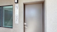 This single-floor space at unit 3107 presents a clean, modern aesthetic from the moment you arrive at its entry. The exterior features a textured stucco facade with a sleek, contemporary front door finished in brushed bronze tones, accented by modern lever hardware and a bold vertical unit number display — setting the tone for the interior design.

Inside, the open-concept layout seamlessly connects the kitchen and living areas, creating a spacious, airy feel enhanced by light hardwood-style flooring throughout. The kitchen is well-appointed with dark espresso cabinetry, a stainless steel refrigerator, a stainless steel range, and a subway tile backsplash. A prominent stainless steel range hood serves as a focal point above the cooking area. The white kitchen island with an integrated sink provides additional prep space and casual seating, bridging the kitchen and living zone.

Recessed lighting and a trio of industrial-style pendant lights illuminate the kitchen, while a ceiling fan with integrated lighting services the adjacent living area. The crisp white walls and bright recessed lighting throughout contribute to a fresh, open ambiance well-suited to contemporary living.
