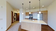 This single-floor space is designed with a clean, contemporary aesthetic that emphasizes functionality and open living. The kitchen serves as the centerpiece, anchored by a generously sized island with a light-toned countertop and an undermount sink, offering ample workspace and a natural gathering point. Three pendant lights with white shades hang above the island, providing focused task lighting while contributing to the modern design.

The kitchen cabinetry features warm wood-tone finishes on both upper and lower units, complemented by a full suite of stainless steel appliances, including a refrigerator, electric range with microwave above, and a built-in microwave. Recessed lighting throughout the ceiling adds to the bright, well-lit atmosphere of the space.

Flooring transitions between hardwood-style material in the kitchen and dining area and carpet in the adjacent living zone, creating a subtle distinction between functional and relaxed areas. A dedicated laundry nook, visible from the main living area, houses a stacked washer and dryer unit, offering convenient in-unit laundry. The overall ambiance is clean, bright, and practical.