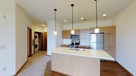 This single-floor space is designed with a clean, contemporary aesthetic that emphasizes functionality and open living. The kitchen serves as the centerpiece, anchored by a generously sized island with a light-toned countertop and an undermount sink, offering ample workspace and a natural gathering point. Three pendant lights with white shades hang above the island, providing focused task lighting while contributing to the modern design.

The kitchen cabinetry features warm wood-tone finishes on both upper and lower units, complemented by a full suite of stainless steel appliances, including a refrigerator, electric range with microwave above, and a built-in microwave. Recessed lighting throughout the ceiling adds to the bright, well-lit atmosphere of the space.

Flooring transitions between hardwood-style material in the kitchen and dining area and carpet in the adjacent living zone, creating a subtle distinction between functional and relaxed areas. A dedicated laundry nook, visible from the main living area, houses a stacked washer and dryer unit, offering convenient in-unit laundry. The overall ambiance is clean, bright, and practical.