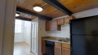 This two-floor space showcases a distinctive blend of rustic character and practical design. The kitchen is a standout feature, highlighted by rich wood-plank ceilings supported by dark painted exposed beams that lend a warm, cabin-like ambiance to the room. Hardwood floors run throughout, adding to the space's timeless aesthetic. The kitchen is equipped with dark black appliances, including a refrigerator, electric range, and dishwasher, complemented by warm wood cabinetry and granite-style countertops that provide ample workspace. A double sink and overhead cabinetry offer functional storage and prep space. Natural light fills the area through multiple windows, brightening the open layout. Louvered bi-fold doors add a classic touch, providing access to additional areas of the space. An adjacent bathroom is visible from the kitchen corridor, featuring a vanity with granite-style countertops and warm wood cabinetry that mirrors the kitchen's design language. Flush-mount ceiling fixtures provide consistent lighting throughout. The overall ambiance is cozy yet functional, with the exposed wood ceiling and hardwood floors creating a cohesive, character-rich environment across both floors of the space.