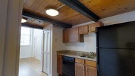 This two-floor space showcases a distinctive blend of rustic character and practical design. The kitchen is a standout feature, highlighted by rich wood-plank ceilings supported by dark painted exposed beams that lend a warm, cabin-like ambiance to the room. Hardwood floors run throughout, adding to the space's timeless aesthetic. The kitchen is equipped with dark black appliances, including a refrigerator, electric range, and dishwasher, complemented by warm wood cabinetry and granite-style countertops that provide ample workspace. A double sink and overhead cabinetry offer functional storage and prep space. Natural light fills the area through multiple windows, brightening the open layout. Louvered bi-fold doors add a classic touch, providing access to additional areas of the space. An adjacent bathroom is visible from the kitchen corridor, featuring a vanity with granite-style countertops and warm wood cabinetry that mirrors the kitchen's design language. Flush-mount ceiling fixtures provide consistent lighting throughout. The overall ambiance is cozy yet functional, with the exposed wood ceiling and hardwood floors creating a cohesive, character-rich environment across both floors of the space.