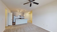 This single-story residence features three bedrooms and two bathrooms within a thoughtfully arranged floor plan. The open-concept living and kitchen area serves as the heart of the home, creating a cohesive and functional shared space. The kitchen is equipped with stainless steel appliances, including a refrigerator and range, and is accented by light wood cabinetry with a tile backsplash and dark stone countertops. A central kitchen island provides additional workspace and casual seating, while pendant lighting above the island adds a decorative touch alongside recessed ceiling lights. The living area features neutral wall-to-wall carpeting that contributes to a warm, comfortable atmosphere, and a ceiling fan overhead promotes air circulation throughout the space. The clean, neutral color palette of white walls and light finishes gives the home a bright, modern feel. The layout flows openly between the kitchen and living space, making it well-suited for everyday living and entertaining. The overall design emphasizes practicality and comfort, with consistent finishes throughout that create a cohesive aesthetic.