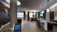 Located at 201 Willett Ave in Port Chester, NY, this single-floor amenity lounge is designed with a sleek, contemporary aesthetic that prioritizes both style and functionality. The open-plan layout creates a generous sense of space, allowing multiple activity zones to coexist seamlessly. Dark hardwood-style flooring runs throughout, grounding the room with a warm yet sophisticated tone that complements the neutral gray and taupe color palette.

At the center of the space, a billiards table serves as a natural focal point, surrounded by ample room for play. To one side, a well-appointed bar counter with tall, cushioned bar stools invites social gathering, accented by pendant lighting that casts a warm, intimate glow. Built-in shelving and cabinetry behind the bar add functional storage while enhancing the polished look of the area.

A lounge seating area anchors another corner of the room, featuring upholstered chairs with accent pillows that introduce a pop of color. Large windows dressed in floor-length drapes allow natural light to filter in, brightening the space while maintaining an air of privacy. Recessed lighting and wall sconces supplement the ambient illumination throughout. A mounted flat-screen television adds an entertainment dimension, making this lounge a versatile and inviting gathering space.