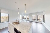 Located on the third floor at 1465 Commonwealth Ave in Boston's Brighton neighborhood, this well-appointed 2-bedroom, 2-bathroom residence offers a thoughtfully designed open-concept layout. Dark hardwood flooring flows throughout, complementing the clean, modern aesthetic of the bright white walls and trim.

The kitchen is a standout feature, boasting marble-veined countertops, white shaker cabinetry, stainless steel appliances — including a range, microwave, refrigerator, and dishwasher — and a central island with pendant lighting that transitions seamlessly into the living area. The spacious living room is lined with large, floor-to-ceiling windows that flood the space with natural light and offer expansive views of the Boston cityscape and Commonwealth Avenue below.

Both bedrooms are well-proportioned and bright, each benefiting from generous windows with city views and ample closet space — five closets are distributed throughout the unit. The two bathrooms are finished with modern gray vanities, clean white countertops, and light wood-toned tile flooring. A hallway connects all areas efficiently, with an additional utility room rounding out the floor plan. This residence combines contemporary finishes with a prime location along one of Boston's most recognized corridors.