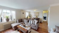 This single-floor residence offers a well-organized layout with distinct living, sleeping, and kitchen areas connected by warm hardwood flooring and a consistent neutral color palette throughout. The expansive living room serves as the centerpiece of the home, featuring rich hardwood floors, crown molding, and large windows that allow natural light to fill the space. A generous sectional sofa anchors one end of the room, while the opposite side provides a comfortable seating nook with an armchair and decorative open shelving near the front entry. The kitchen is efficiently designed with white cabinetry, dark countertops, a tile backsplash, and tile flooring, offering ample storage and workspace. The residence includes multiple bedrooms, each carpeted and equipped with ceiling fans and radiator heating. The primary bedroom features a window unit and natural light, while additional bedrooms provide flexible use as sleeping quarters or hobby spaces. Each bedroom is finished with neutral walls, offering a calm and restful environment. Throughout the home, consistent design choices—including classic trim work, soft tones, and curated wall art—contribute to a cohesive and inviting atmosphere.