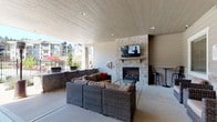 This covered outdoor patio is designed as a versatile communal lounge space, offering a well-appointed environment suitable for use across multiple seasons. The area is sheltered beneath a wood-paneled ceiling with recessed lighting, providing a sense of enclosure while remaining open to the surrounding outdoors. Stone columns frame the perimeter, reinforcing the space's architectural character and complementing the standout stacked-stone fireplace, which serves as the focal point of the main seating zone. A flat-screen television is mounted above the fireplace's wood mantel, making the area well-suited for both relaxed gatherings and viewing events.

The seating is arranged thoughtfully throughout the patio. A large wicker sectional sofa with cushioned upholstery is positioned facing the fireplace, accompanied by a glass-top coffee table. Additional wicker lounge chairs and smaller bistro-style high-top tables provide supplemental seating options. Freestanding patio heaters are positioned throughout, extending the usability of the space during cooler temperatures. A dining table with chairs occupies a separate section near a glass entry door and window, offering a more structured seating alternative. The concrete flooring and neutral, earth-toned palette throughout unify the space with a clean, contemporary aesthetic.