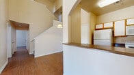 This two-story home offers four bedrooms and two bathrooms distributed across a well-organized floor plan. The main floor provides the larger share of the living space and features an open-concept layout connecting the living area, kitchen, and dining space. The kitchen includes a breakfast bar with dark countertops, white cabinetry, and a full-size refrigerator, with an arched pass-through that adds architectural character. A pendant light fixture hangs above the bar, complementing the warm hardwood-style flooring found throughout the home.

The living room is a standout space, featuring soaring vaulted ceilings, a ceiling fan, and a sliding glass door that opens to a private balcony with outdoor views — filling the room with natural light. The staircase connects to the upper floor, which houses additional bedrooms, including the generously sized primary bedroom.

The primary bedroom features its own vanity area with a wide mirror, dark countertop, and ample cabinet storage, along with a dedicated walk-in closet equipped with wire shelving. The bathrooms include a full tub-shower combination. Additional closets with wire shelving systems provide practical storage throughout the home.