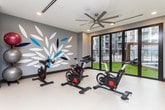 This single-floor fitness studio is thoughtfully designed to support a well-rounded workout experience. The space is anchored by a row of spin bikes positioned to take full advantage of the floor-to-ceiling glass doors and expansive windows along one wall, which flood the room with natural light and provide views of an outdoor pool and courtyard area beyond. A large wall-mounted mirror runs the length of one side, visually expanding the space and supporting form-conscious training, while a mounted flat-screen display offers on-demand fitness content. The flooring throughout is a smooth, durable light-toned surface well-suited for fitness use. A decorative botanical mural featuring white and blue leaf motifs adds a stylish, contemporary touch to the grey-toned walls. The studio is equipped with a full rack of dumbbells, stability balls, and built-in storage cubbies for accessories, ensuring that users have a broad range of training options. A ceiling fan provides consistent air circulation across the open floor, which offers ample room for stretching or group exercise. Recessed lighting and a clean, organized layout contribute to the space's polished and functional ambiance, making it an inviting environment for cardio, strength, and flexibility training alike.