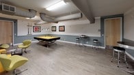 This single-floor community amenity space is designed for recreation and social gathering, blending an industrial aesthetic with bold, contemporary accents. The main area is anchored by a striking pool table with a bright yellow felt surface, suspended beneath exposed ductwork that runs along the ceiling, lending the space an urban, loft-like character. Light wood-toned luxury vinyl flooring extends throughout, complementing the two-tone gray wall treatment that defines the room's color palette.

A dedicated lounge area features a collection of modern yellow pod-style chairs and matching cubic ottomans arranged around a circular glass coffee table, creating an inviting spot for conversation. Colorful framed artwork adorns the walls, adding visual interest and personality to the space.

Along one wall, a set of sleek chrome bistro tables and bar stools provide additional casual seating. The room also includes a well-appointed kitchenette with light maple cabinetry, dark laminate countertops, a stainless steel sink, and a full-size refrigerator — ideal for entertaining. Overhead fluorescent lighting ensures the space remains bright and functional. Together, these elements create a lively, well-equipped amenity room suited for both relaxed socializing and active recreation.