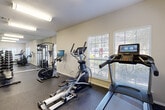 This fitness center is a thoughtfully designed, single-floor space built to support a wide range of workout routines. The room features durable speckled rubber flooring throughout, providing a safe and practical surface for exercise. A full-length mirrored wall runs along one side, visually expanding the space and allowing users to monitor their form during workouts.

Cardio equipment is well-represented, with a treadmill, elliptical trainer, and a stationary spin bike — the latter equipped with an integrated display screen for guided workout sessions. For strength training, the space includes a multi-functional cable weight machine and a dedicated dumbbell rack stocked with a comprehensive range of free weights, accompanied by a adjustable workout bench.

A separate accessory station along one wall holds an assortment of stability and exercise balls in multiple sizes, along with resistance bands and foam rollers, catering to flexibility and functional training needs.

Natural light filters into the room through multiple windows fitted with white horizontal blinds, offering views of surrounding greenery. Overhead fluorescent lighting ensures the space remains well-lit at all times. The neutral, light-toned walls and textured ceiling contribute to a clean, focused environment. A door provides internal access, suggesting the fitness center is part of a larger facility.