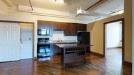 This single-floor loft-style space showcases a thoughtfully designed open layout that blends industrial character with modern comfort. The kitchen serves as a central highlight, featuring rich dark wood cabinetry, light stone countertops, and a full suite of black appliances including a gas range, microwave, dishwasher, and refrigerator. A freestanding kitchen island with a built-in wine rack adds both functionality and style, making it an ideal space for cooking and entertaining. The kitchen flows seamlessly into the living area, enhancing the sense of openness throughout. Underfoot, wide-plank hardwood floors in a deep, warm tone lend a sense of character and warmth to the space. Overhead, exposed ductwork and industrial-style track lighting reinforce the loft aesthetic while providing excellent illumination. The elevated ceilings amplify the feeling of spaciousness, creating an airy and open ambiance. The warm, neutral-toned walls complement the dark architectural elements, striking a balance between cozy and contemporary. Overall, this space offers a distinctive urban living experience rooted in industrial design with practical, well-appointed features throughout.