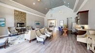 Located at Arbor Keats Drive in Simpsonville, South Carolina, this single-floor community space offers a thoughtfully designed layout that seamlessly combines a generous living area with a functional kitchen. The living room is the architectural centerpiece, boasting dramatic vaulted ceilings with recessed lighting that amplify the sense of openness and volume. Warm hardwood-style flooring runs throughout, providing a cohesive foundation for the space. Large French doors and tall arched windows flood the interior with natural light while offering views of the surrounding outdoors, enhanced by elegant drapery panels. The living area is furnished with multiple neutral-toned accent chairs, a tufted ottoman, and curated artwork, creating an inviting and sophisticated lounge atmosphere. A stacked-stone fireplace accent wall adds texture and warmth to one end of the room. The kitchen is well-appointed with rich dark wood cabinetry, granite countertops, stainless steel appliances including a French door refrigerator and dishwasher, and a wall-mounted television. A sturdy farmhouse-style dining table with upholstered seating bridges the kitchen and living areas, making the space well-suited for both casual gatherings and community events.