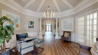 Located at 100 Ivey Park Ln in Norcross, Georgia, this sophisticated single-floor space exudes a refined, classical aesthetic defined by thoughtful architectural detailing and a welcoming ambiance. The centerpiece of the interior is a striking octagonal reception area crowned by a soaring coffered ceiling with crisp white trim and a dramatic geometric chandelier in brushed gold, casting warm light across rich hardwood flooring that extends throughout the space. Stately decorative columns frame the transition between areas, reinforcing the formal yet approachable character of the interior. Wainscoting and panel molding line the walls in a neutral warm-gray palette, lending a polished, cohesive look to every room. A tastefully appointed entry foyer features a gold-framed mirror, a marble-top console table, and soft accent lighting, creating an inviting first impression. The main workspace is equipped with a modern desk and seating area suited for client consultations, complemented by upholstered accent chairs and abundant natural light streaming through French doors and large windows. A secondary gathering room opens off the main corridor, featuring French doors, drapery panels, and casual dining-style seating bathed in natural light. Throughout, carefully selected artwork and greenery add warmth and character to this elegantly designed professional environment.