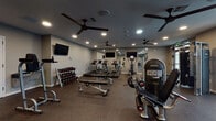 Located at 505 W 7th St in New Castle, Delaware, this single-level fitness center offers a comprehensive workout environment designed to support a wide range of exercise routines. The space is organized into distinct functional zones, providing users with dedicated areas for cardio, strength training, and functional fitness.

The cardio section features a row of treadmills positioned alongside a mirrored wall, enhancing the sense of depth and allowing users to monitor their form. Wall-mounted flat-screen televisions are positioned throughout to provide entertainment during workouts. Ceiling fans are distributed across the facility, contributing to airflow and comfort during exercise.

The strength training area is equipped with multiple selectorized weight machines, including leg extension and cable pulley systems, catering to both upper and lower body workouts. A well-stocked dumbbell rack offers a full range of free weights, supported by a flat bench for versatile use.

The functional training zone includes a dedicated rack of stability balls, resistance bands, medicine balls, and balance tools, making it suitable for dynamic and rehabilitative exercises. Large windows along one wall allow natural light to fill the space, creating an open and energizing atmosphere. Rubber flooring throughout provides durability, safety, and noise reduction. Access is provided through glass double doors, connecting the fitness center to adjacent common areas.