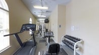 This fitness room offers a comprehensive workout environment designed to accommodate a range of exercise needs. The space is laid out in a narrow, elongated floor plan that makes efficient use of its footprint. Ceiling fans are evenly spaced along the length of the room, ensuring adequate air circulation during workouts, while flush-mounted fluorescent light fixtures provide consistent overhead illumination throughout.

On the left side of the room, cardio equipment is positioned along the wall, including at least one treadmill with a digital console. Toward the center of the room, a multi-station cable and weight machine offers versatile strength training options. A stationary recumbent-style bike is also present near the middle of the space, providing a low-impact cardio option. Along the right wall, a well-organized dumbbell rack holds a graduated selection of free weights, catering to users at various fitness levels.

The walls are finished in a neutral, warm beige tone, contributing to a clean and welcoming atmosphere. A wall-mounted television is positioned mid-room, visible from multiple workout stations. Natural light enters through a window at the far end of the room, adding a bright, airy quality to the space.