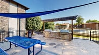 This outdoor communal patio space is thoughtfully designed for social gatherings and al fresco dining. The centerpiece of the area is a built-in outdoor kitchen station, constructed with a stacked stone surround and equipped with multiple stainless steel gas grills, providing ample cooking capacity for group events. A classic wooden pergola frames the grilling station, adding a structured architectural element while allowing open-air enjoyment of the surrounding greenery.

Vibrant blue shade sails are strategically positioned overhead, offering relief from direct sunlight across the concrete patio surface. Matching blue steel mesh picnic tables with attached bench seating provide comfortable dining and gathering spaces beneath the shaded area. A decorative metal railing encloses the perimeter of the patio, delineating the space while maintaining open sightlines to the manicured lawn and mature trees beyond.

The surrounding landscape features lush green grass and well-established deciduous trees, contributing to a pleasant, park-like atmosphere. The brick exterior of the adjacent building complements the stone detailing of the grill station, tying the overall aesthetic together cohesively. A pool area is visible in the background, suggesting this patio serves as part of a larger outdoor amenity complex within the community.