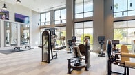 Located at 5919 Ashlar Way in Alexandria, VA, this contemporary fitness center offers a well-appointed, single-floor layout divided into two distinct areas: a main workout floor and a dedicated studio space. The main gym is defined by dramatically tall ceilings and expansive floor-to-ceiling windows that flood the space with natural light and provide open views of the surrounding greenery. The area is equipped with a comprehensive selection of strength machines, cable systems, treadmills, ellipticals, stationary bikes, rowing machines, and a dedicated free weights section featuring dumbbell racks and a flat bench. A wall-mounted rack holds an assortment of kettlebells and TRX suspension trainers for functional training. Large mirrors and mounted flat-screen televisions are positioned throughout, while stylish pendant lighting with black fixtures complements the modern, neutral aesthetic of the patterned carpet tile flooring and white brick accent walls. The secondary studio space features light wood flooring, soft green accent walls, and built-in storage cubbies stocked with yoga mats and exercise balls. A wall-mounted display screen and full-length mirrors complete the studio, making it well-suited for stretching, yoga, or guided workouts. Together, these two areas provide a comprehensive and inviting fitness environment.