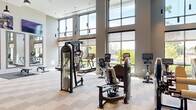 Located at 5919 Ashlar Way in Alexandria, VA, this contemporary fitness center offers a well-appointed, single-floor layout divided into two distinct areas: a main workout floor and a dedicated studio space. The main gym is defined by dramatically tall ceilings and expansive floor-to-ceiling windows that flood the space with natural light and provide open views of the surrounding greenery. The area is equipped with a comprehensive selection of strength machines, cable systems, treadmills, ellipticals, stationary bikes, rowing machines, and a dedicated free weights section featuring dumbbell racks and a flat bench. A wall-mounted rack holds an assortment of kettlebells and TRX suspension trainers for functional training. Large mirrors and mounted flat-screen televisions are positioned throughout, while stylish pendant lighting with black fixtures complements the modern, neutral aesthetic of the patterned carpet tile flooring and white brick accent walls. The secondary studio space features light wood flooring, soft green accent walls, and built-in storage cubbies stocked with yoga mats and exercise balls. A wall-mounted display screen and full-length mirrors complete the studio, making it well-suited for stretching, yoga, or guided workouts. Together, these two areas provide a comprehensive and inviting fitness environment.