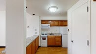 This single-floor unit offers a practical and well-organized layout designed for comfortable everyday living. The kitchen is a standout feature, equipped with warm honey-toned wood cabinetry, granite-style countertops, a full suite of white appliances including a refrigerator, electric range, over-the-range microwave, and a single-basin sink. An additional countertop peninsula provides extra prep space and a natural transition into the adjacent living area. Throughout the unit, light hardwood flooring creates a cohesive and warm aesthetic. The main living area is generously sized, featuring a textured ceiling and a large sliding glass door that invites abundant natural light and provides access to an outdoor space. A secondary room features a window with a curtain, painted cinder block walls, and the same hardwood flooring, lending it a clean, versatile character. The bathroom includes a full soaking tub with a shower, a curtain rod, grab bar safety features, and a warm wood vanity. The overall ambiance of the unit is clean, neutral, and functional, with consistent finishes throughout that create a unified and practical living environment.